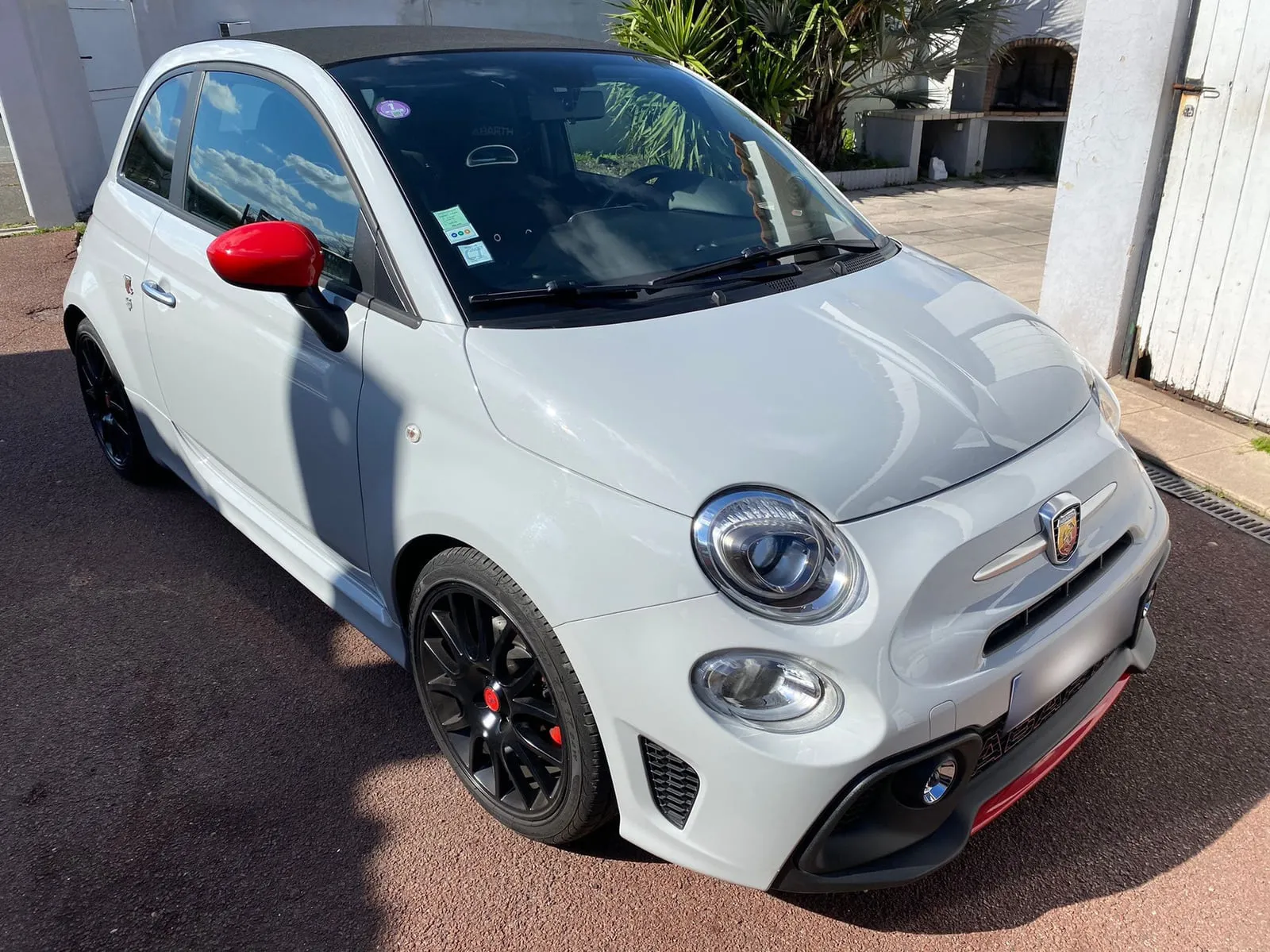 Abarth grise vue de trois quarts avant dans une cour, toit en toile noire : blason Abarth sur le museau, coque de rétroviseur rouge, jantes noires à étriers rouges, lame rouge sous le pare-chocs