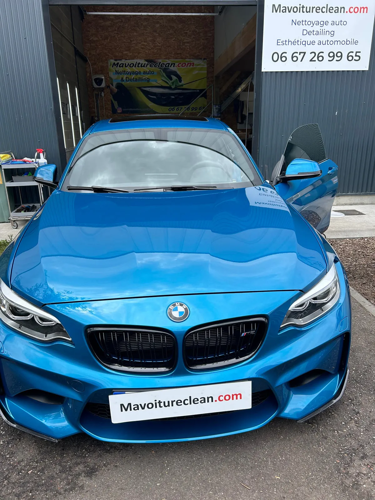 BMW bleue vue de face devant l'atelier, portière ouverte, enseigne Mavoitureclean.com sur la façade