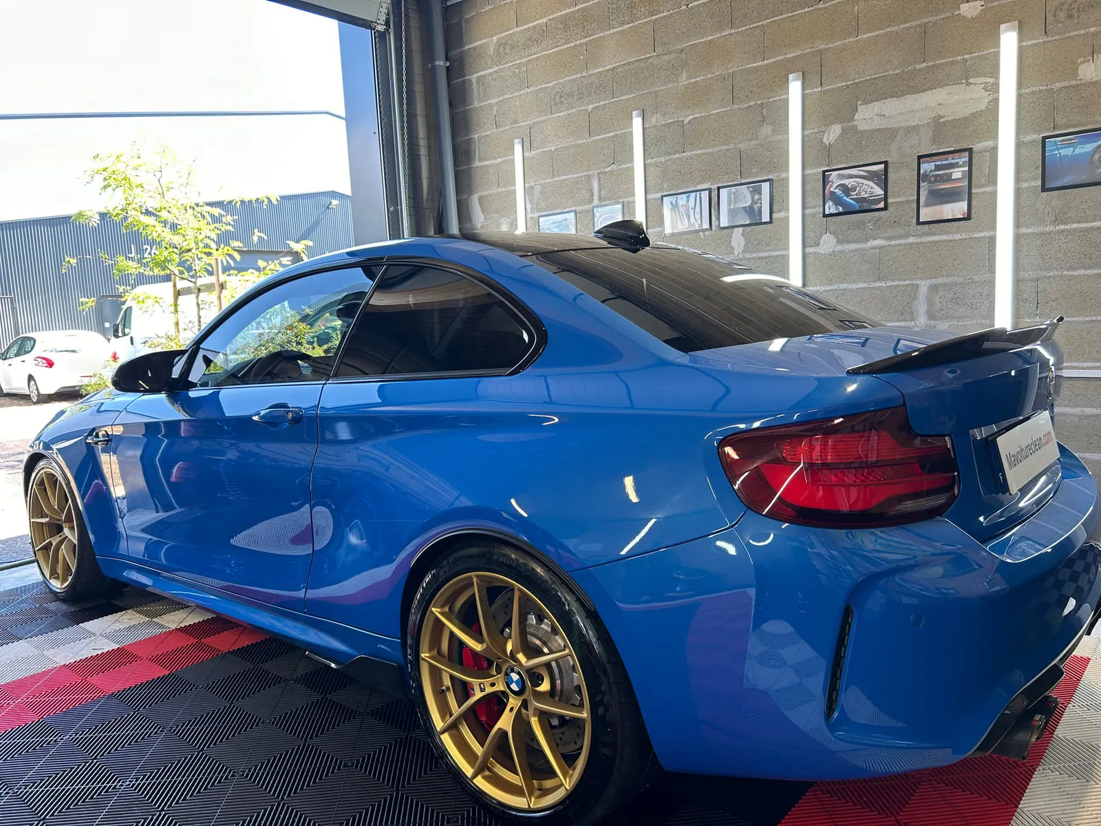 BMW bleue vue de trois quarts arrière dans l'atelier : coupé à aileron noir, jantes dorées à branches fines, étrier rouge, sol rouge et noir, panneau Mavoitureclean.com à la place de la plaque