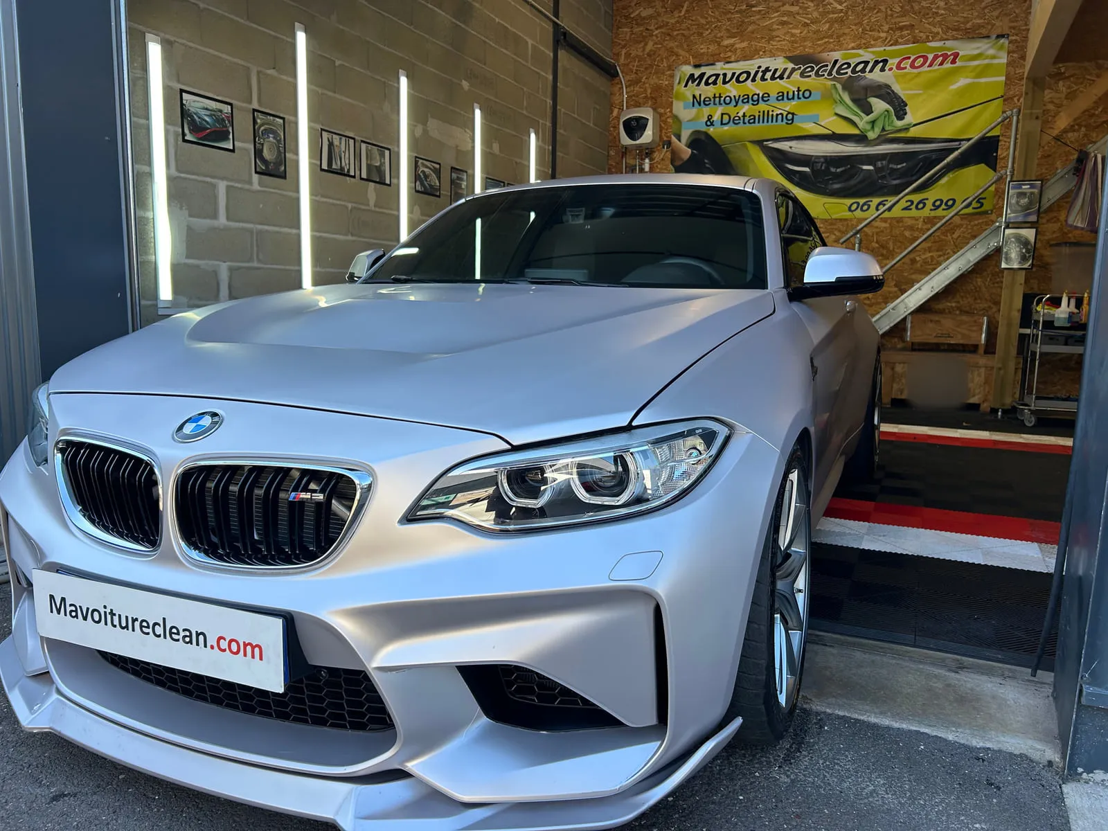 BMW gris mat vue de trois quarts avant à l'entrée de l'atelier, bannière Mavoitureclean.com au fond