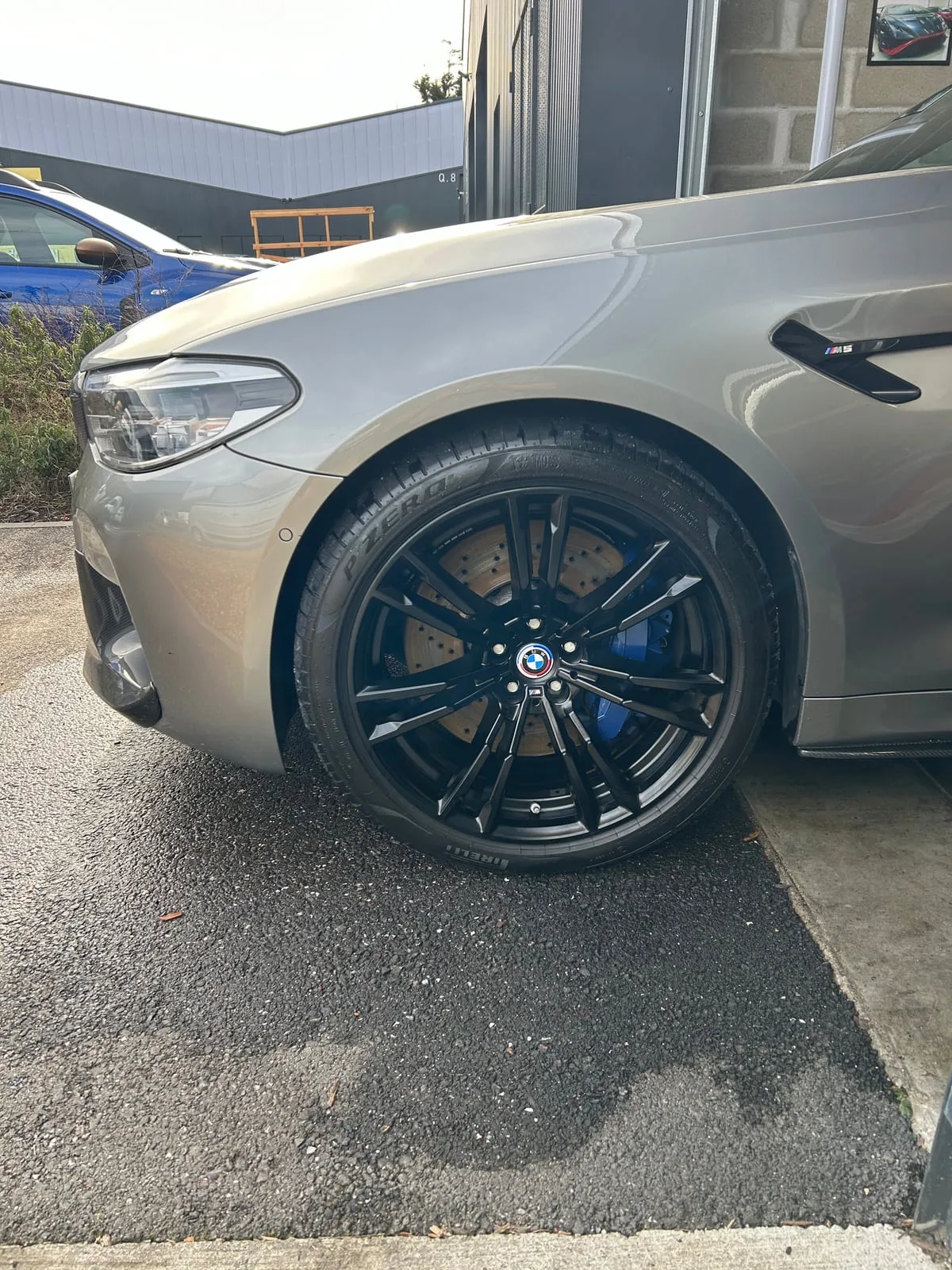 Roue avant d'une BMW M5 grise : jante noire à doubles branches, cache de moyeu BMW, étrier de frein bleu, disque de frein, pneu Pirelli P Zero, badge M5 sur l'aile