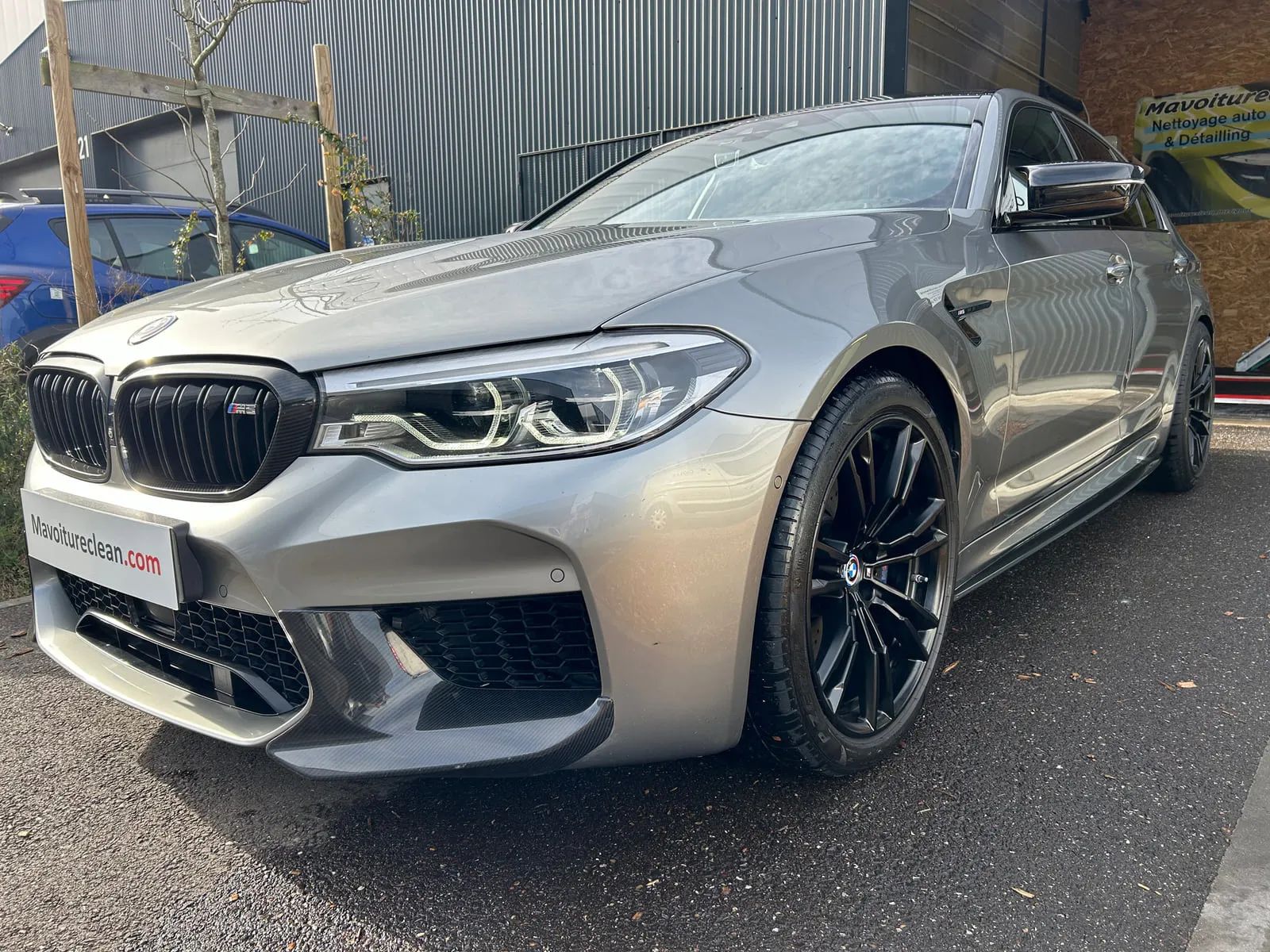BMW grise vue de trois quarts avant devant l'atelier : calandre noire à badge M5, optiques à LED, jantes noires, panneau Mavoitureclean.com à la place de la plaque