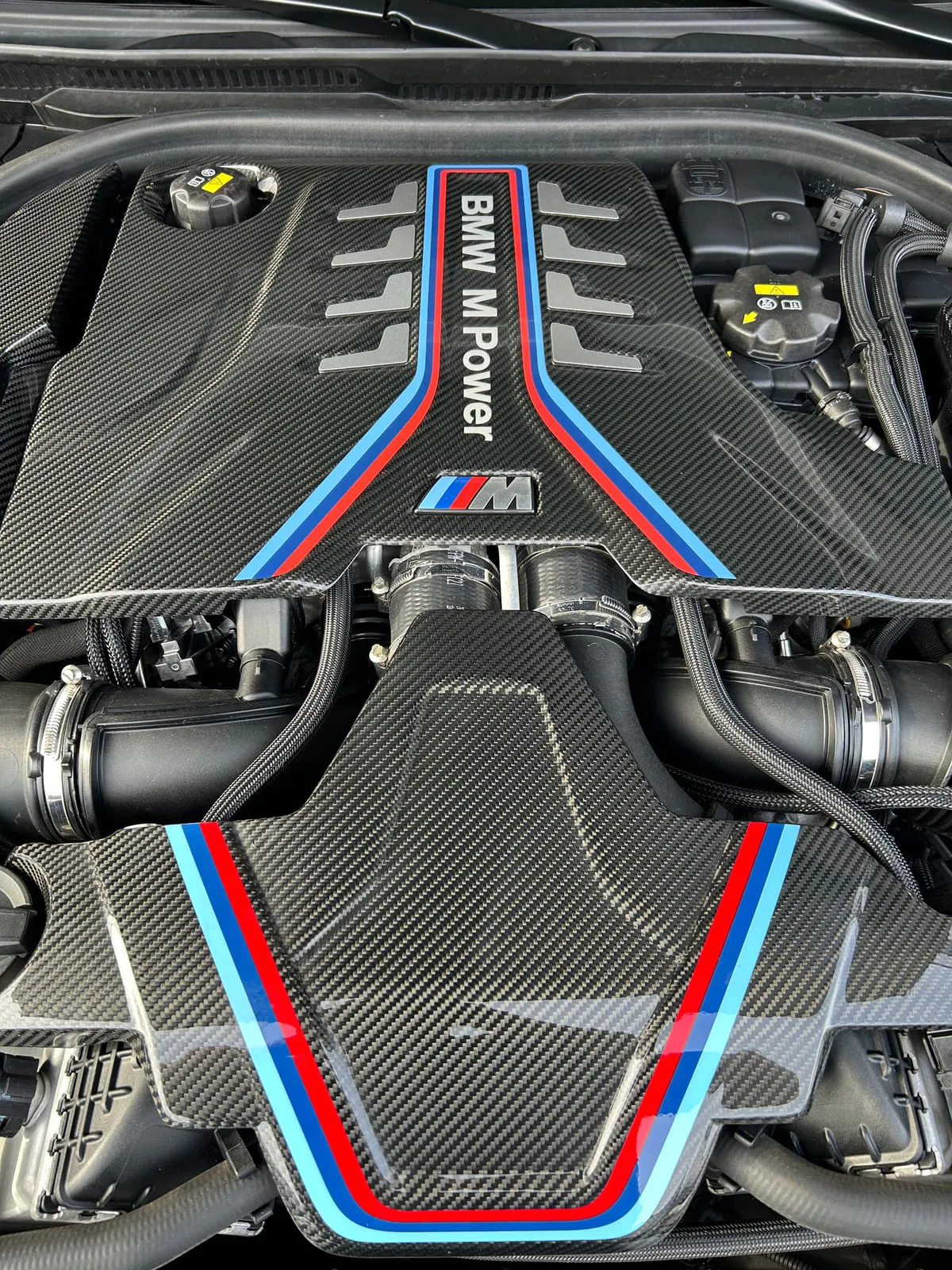 Compartiment moteur d'une BMW capot ouvert : cache moteur en carbone à bandes bleu clair, bleu foncé et rouge, inscription BMW M Power, logo M