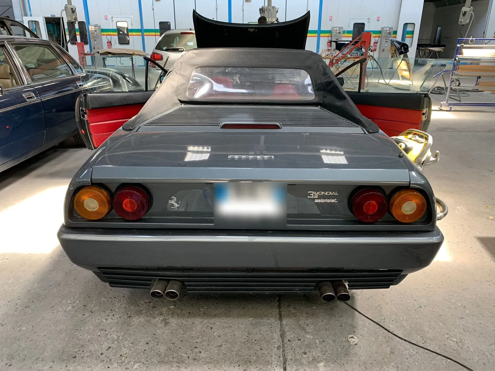 Ferrari grise vue de l'arrière dans un atelier de carrosserie, capote en toile noire fermée et portières ouvertes sur un habitacle rouge : cheval cabré à gauche, inscription Ferrari sur le coffre, badge 3.2 Mondial cabriolet, quatre sorties d'échappement