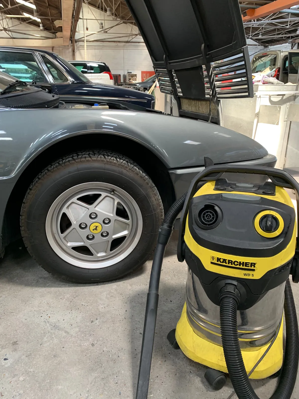 Roue avant d'une Ferrari grise : jante alu à cinq branches, cache de moyeu jaune au cheval cabré, aspirateur eau et poussière Kärcher WD 5 au premier plan, atelier de carrosserie derrière