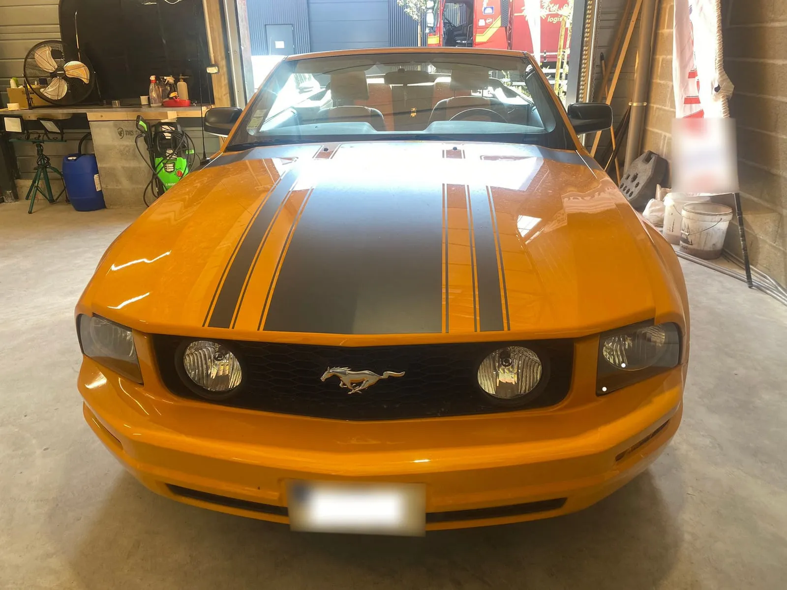 Ford Mustang cabriolet orange vue de face dans un atelier : capot à larges bandes noires, calandre noire au cheval chromé, deux antibrouillards ronds