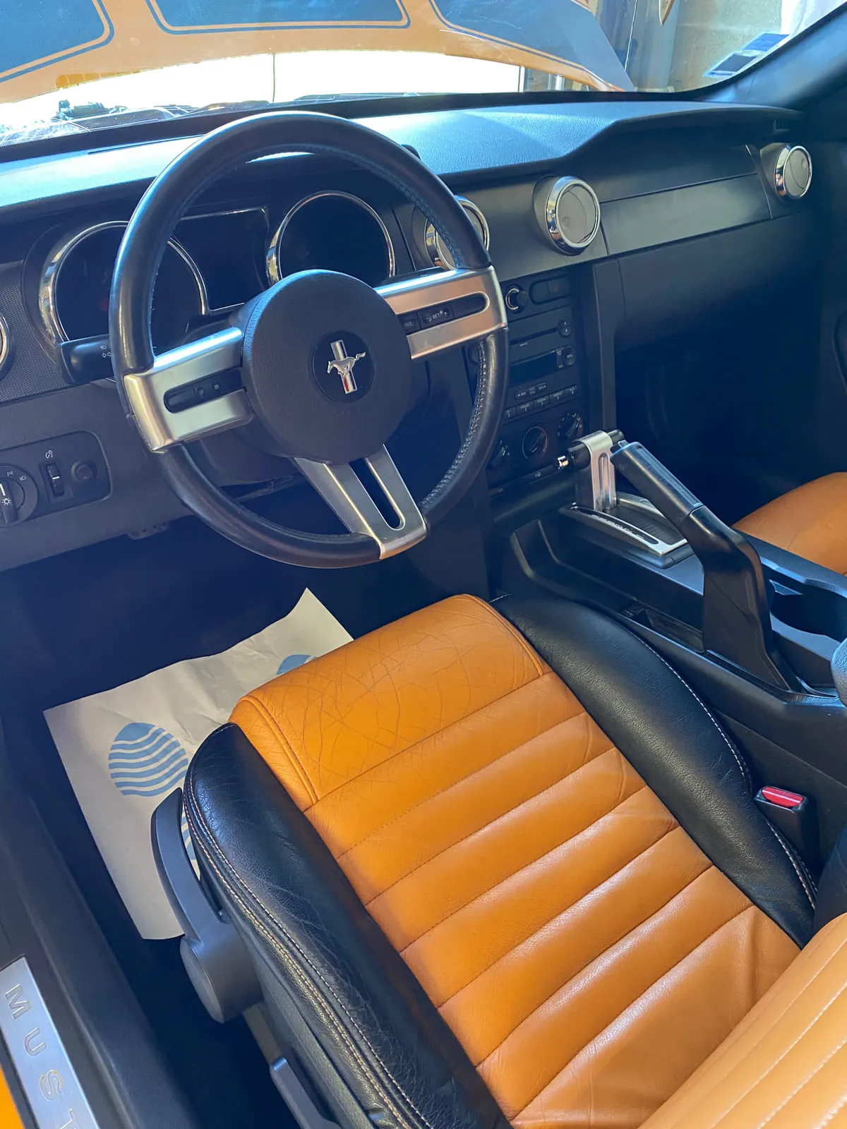 Habitacle d'une Ford Mustang vu par la portière ouverte : volant au cheval Mustang, compteurs cerclés d'argent, levier de boîte automatique, siège en cuir orange et noir, seuil de porte marqué Mustang, feuille de protection en papier sur le plancher
