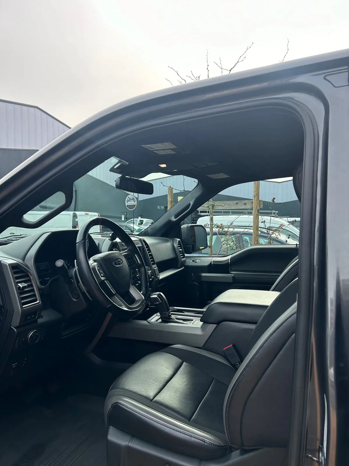 Habitacle d'un Ford vu par la portière avant ouverte : volant à l'ovale Ford, console centrale à levier automatique, sièges en cuir noir perforé, ciel de toit sombre
