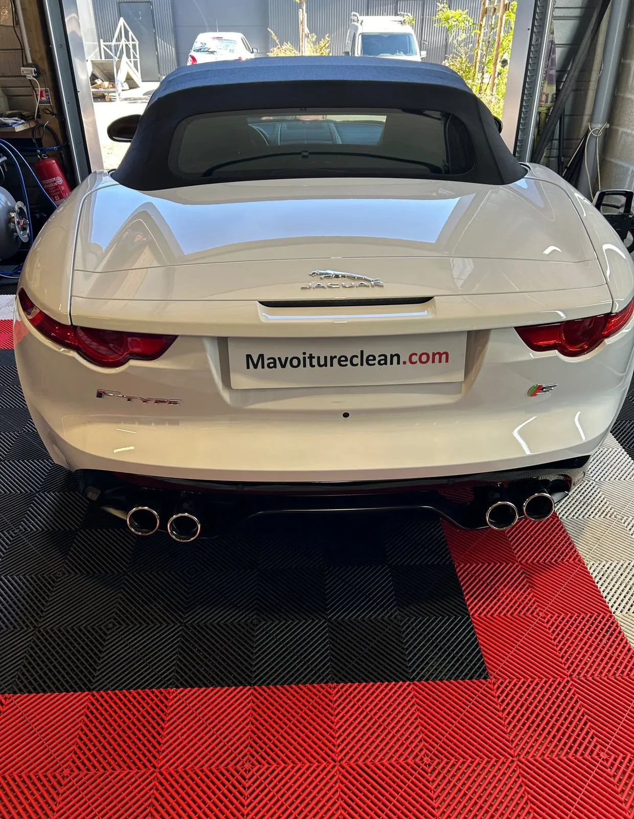 Jaguar F-Type blanche vue de l'arrière dans l'atelier, capote en toile bleu nuit fermée : logo Jaguar sur le coffre, inscription F-TYPE, badge S, quatre sorties d'échappement, panneau Mavoitureclean.com à la place de la plaque