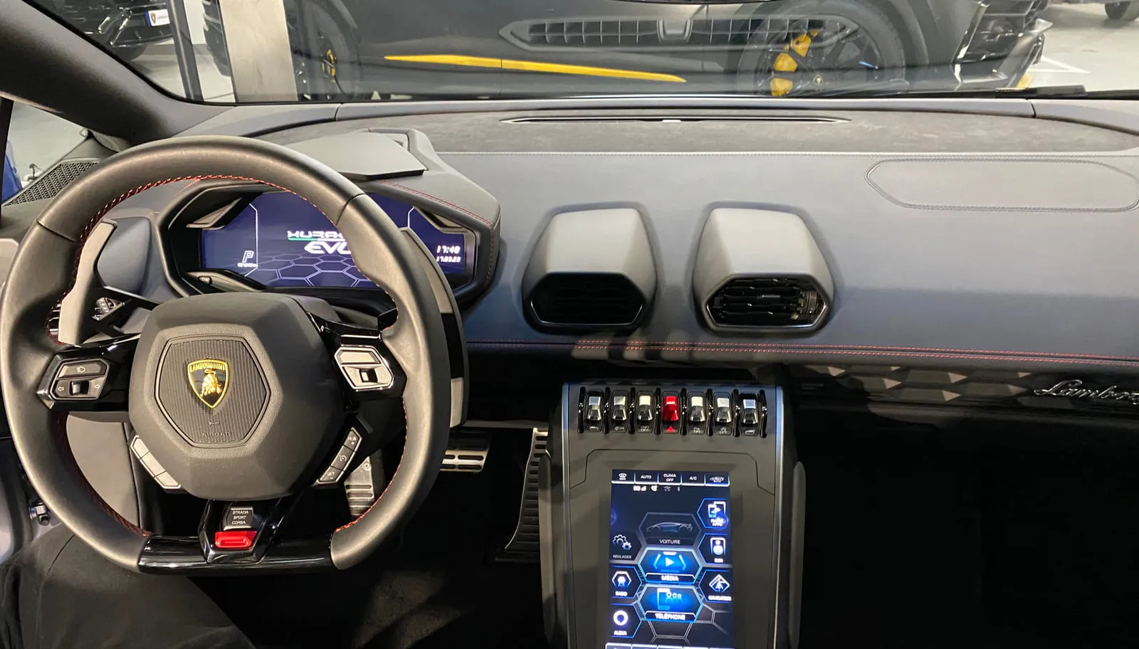 Poste de conduite d'une Lamborghini vu depuis le siège conducteur : volant au blason Lamborghini et aux coutures rouges, combiné numérique qui affiche EVO, écran tactile vertical au centre, rangée de commutateurs, aérateurs hexagonaux