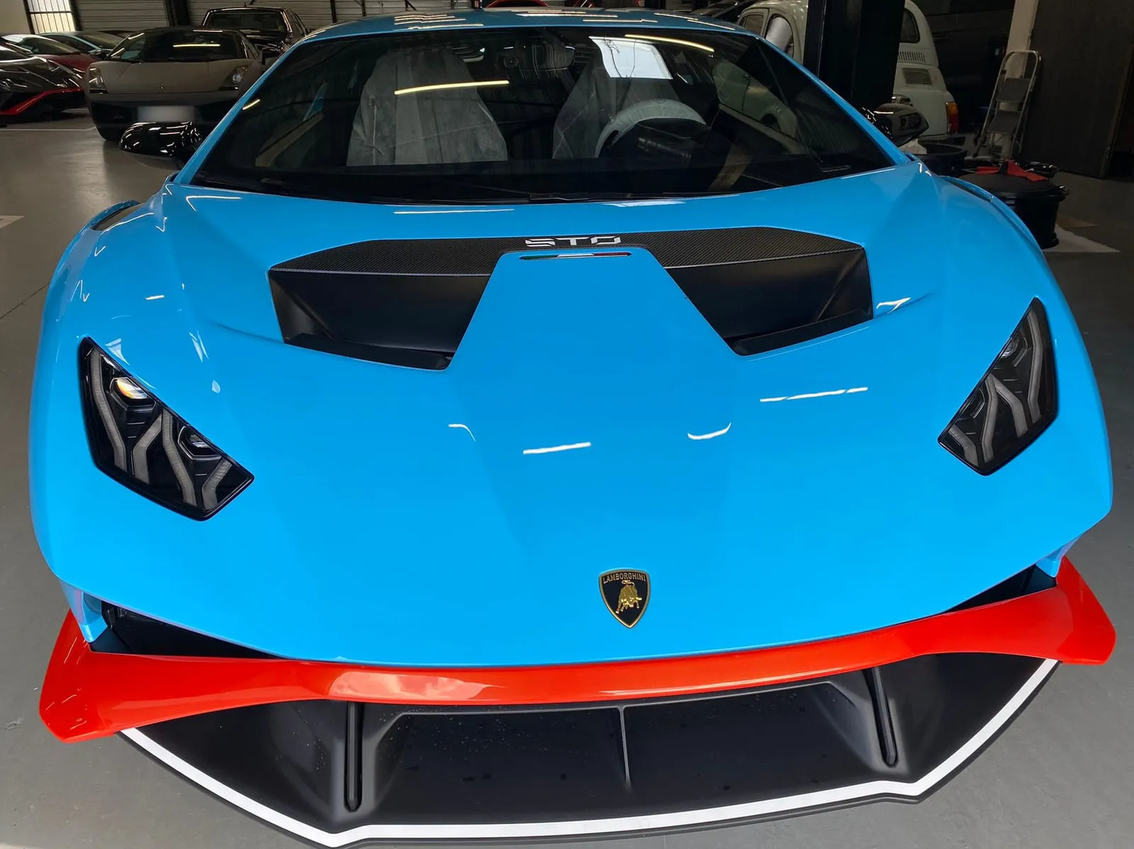 Lamborghini bleu clair vue de face dans un hall où d'autres voitures sont garées : capot à prise d'air en carbone portant l'inscription STO, blason Lamborghini, optiques effilées, grande lame avant rouge, housses en plastique sur les sièges