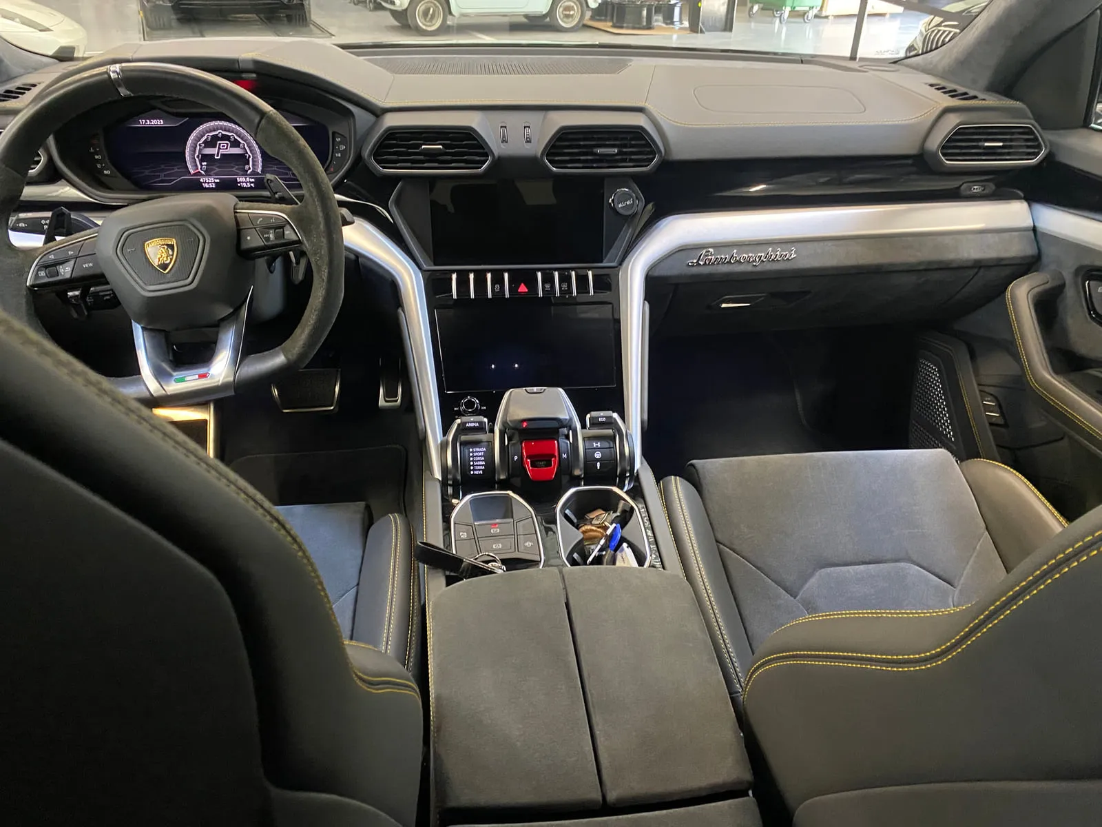 Habitacle d'une Lamborghini vu de l'arrière, entre les dossiers des deux sièges avant : volant au blason Lamborghini et aux surfaces façon daim, deux écrans centraux, montants de console en métal brossé, bouton de démarrage rouge, sièges noirs à surpiqûres jaunes