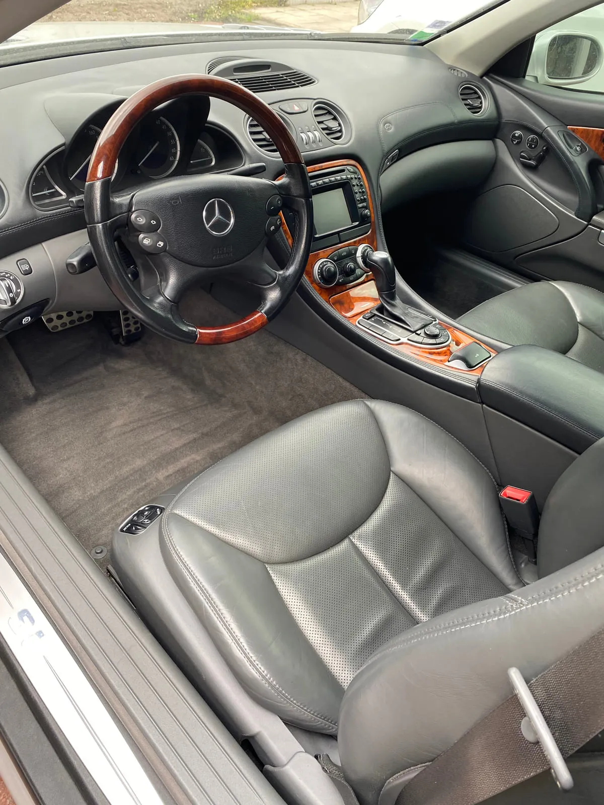 Habitacle d'une Mercedes vu par la portière avant ouverte : volant bois et cuir noir à l'étoile Mercedes, console centrale à décor bois et écran de navigation, siège en cuir gris perforé, moquette grise