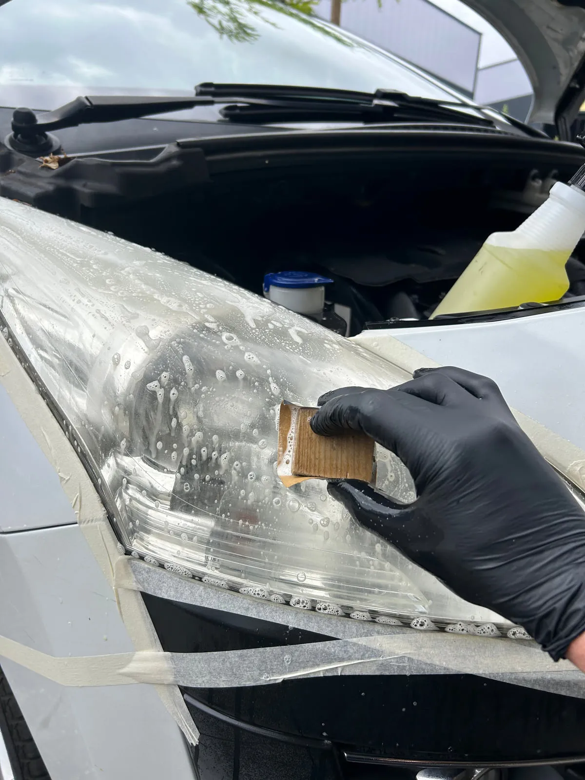 Main gantée de noir frottant avec un abrasif l'optique d'un phare, ruban de masquage tout autour