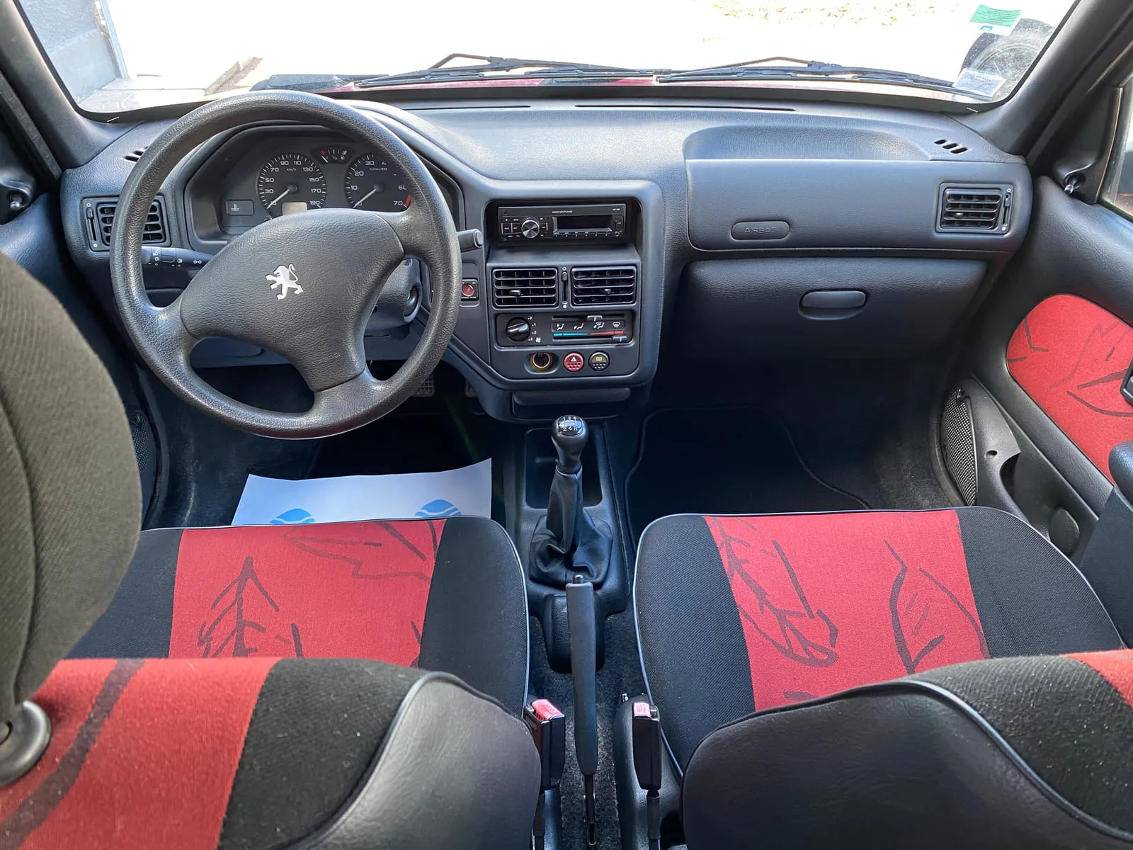 Habitacle d'une Peugeot vu de l'arrière, les deux sièges avant au premier plan : volant au lion Peugeot, deux compteurs ronds, autoradio, levier de vitesses, sièges en tissu rouge et noir à motif de feuilles, feuille de protection en papier posée sur le plancher
