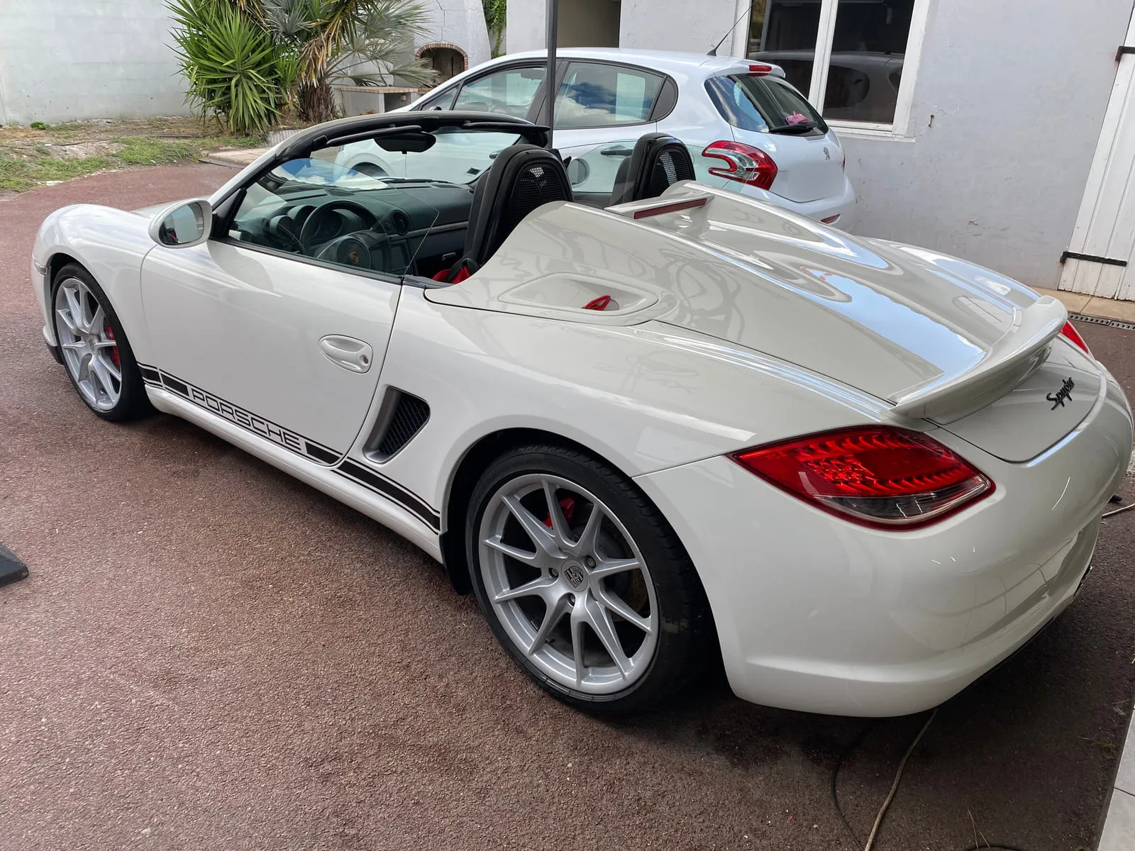 Porsche blanche décapotée vue de trois quarts arrière dans une cour : bandes noires Porsche sur le bas de caisse, inscription Spyder sur la poupe, aileron en bec de canard, jantes argent à rayons fins, étriers rouges