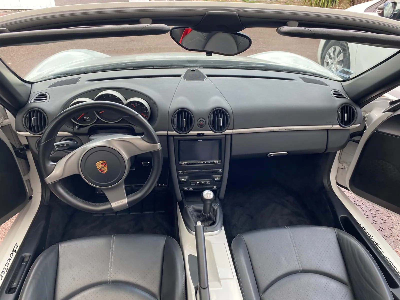 Poste de conduite d'une Porsche Boxster Spyder vu du dessus, toit ouvert : volant au blason Porsche, trois compteurs cerclés d'argent, autoradio, levier de vitesses manuel, sièges en cuir noir, inscription Boxster sur le seuil