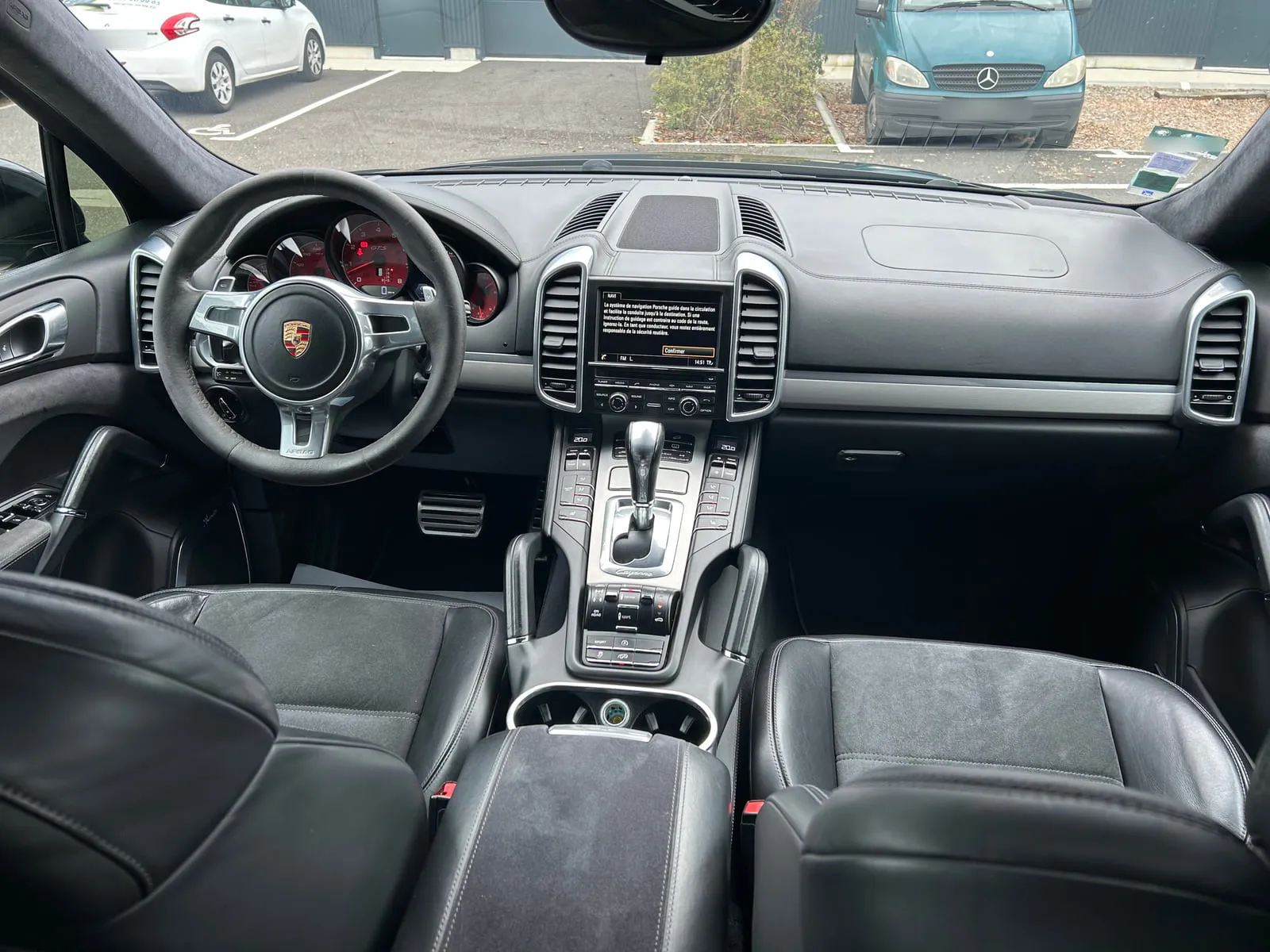 Habitacle d'une Porsche Cayenne vu depuis la banquette arrière : volant au blason Porsche, écran de navigation, aérateurs verticaux, console centrale à levier automatique, sièges avant noirs en cuir et surfaces façon daim