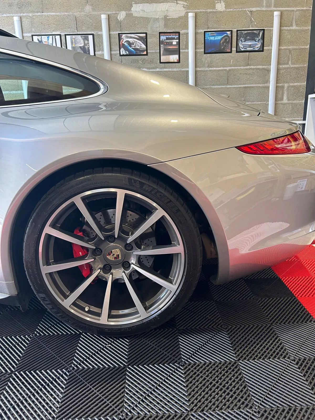 Roue arrière d'une Porsche gris argent : jante bicolore à branches fines, étrier rouge, cache de moyeu au blason Porsche, pneu Michelin, sol d'atelier rouge et noir
