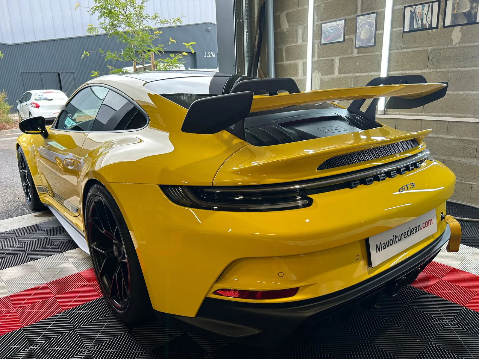 Porsche jaune vue de trois quarts arrière dans l'atelier : grand aileron fixe, inscriptions Porsche et GT3 sur la poupe, sangle jaune au pare-chocs, jantes noires, sol rouge et noir, panneau Mavoitureclean.com à la place de la plaque