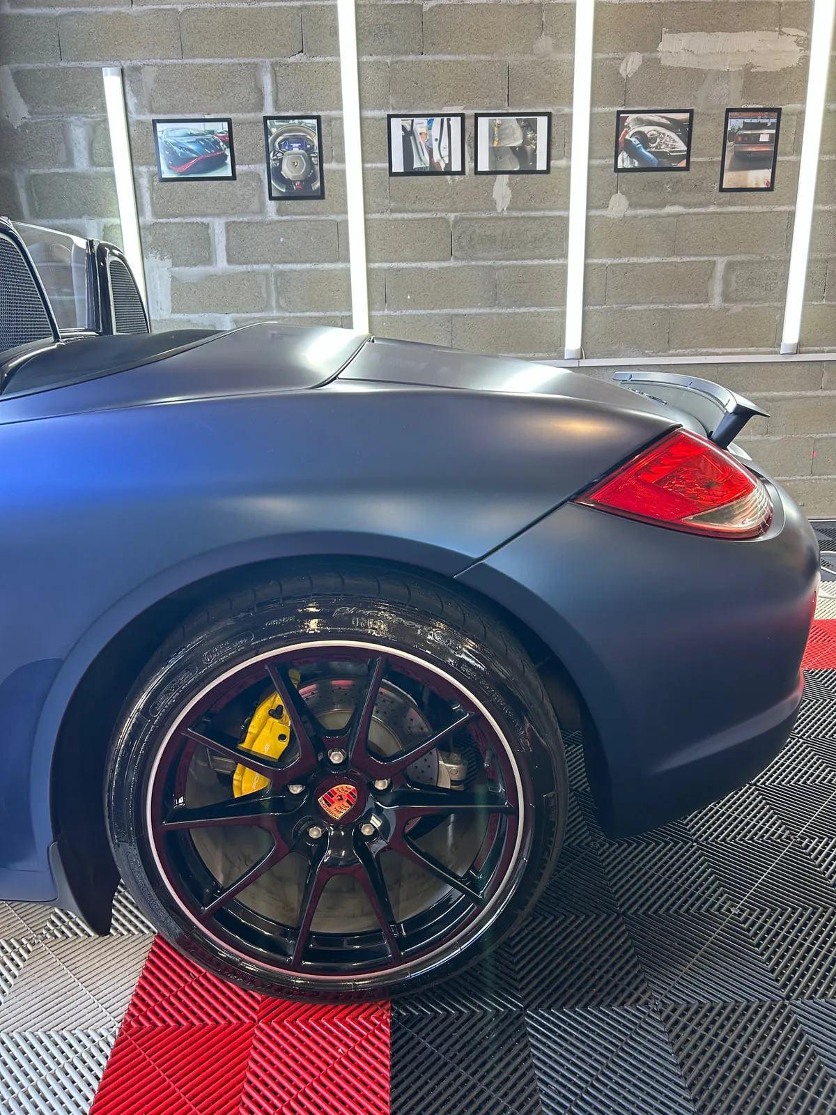 Roue arrière d'une Porsche : jante noire à liseré blanc, étrier jaune, aile bleu mat, sol d'atelier rouge et noir