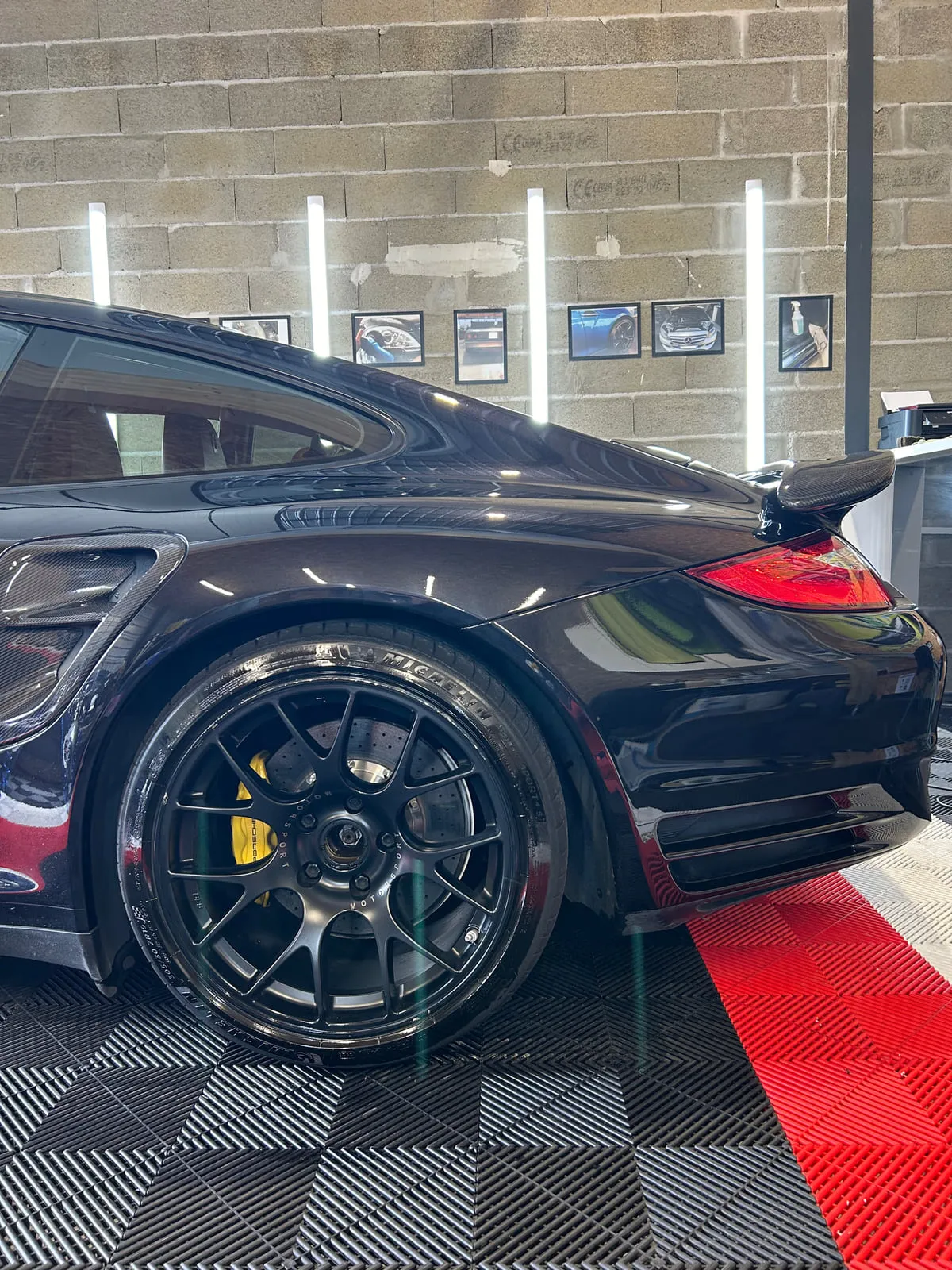 Roue arrière d'une Porsche noire dans l'atelier : jante noire mate à rayons fins, étrier jaune, pneu Michelin, sol rouge et noir