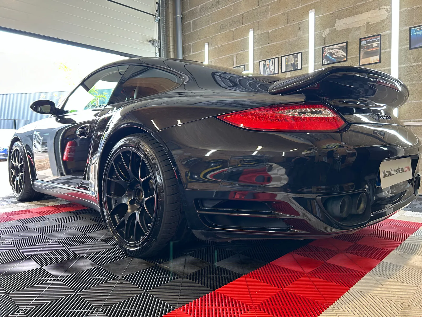 Porsche noire vue de trois quarts arrière dans l'atelier : aileron en carbone, inscription turbo à droite du capot arrière, feux rouges, jantes noires à rayons fins, sol rouge et noir, panneau Mavoitureclean.com à la place de la plaque