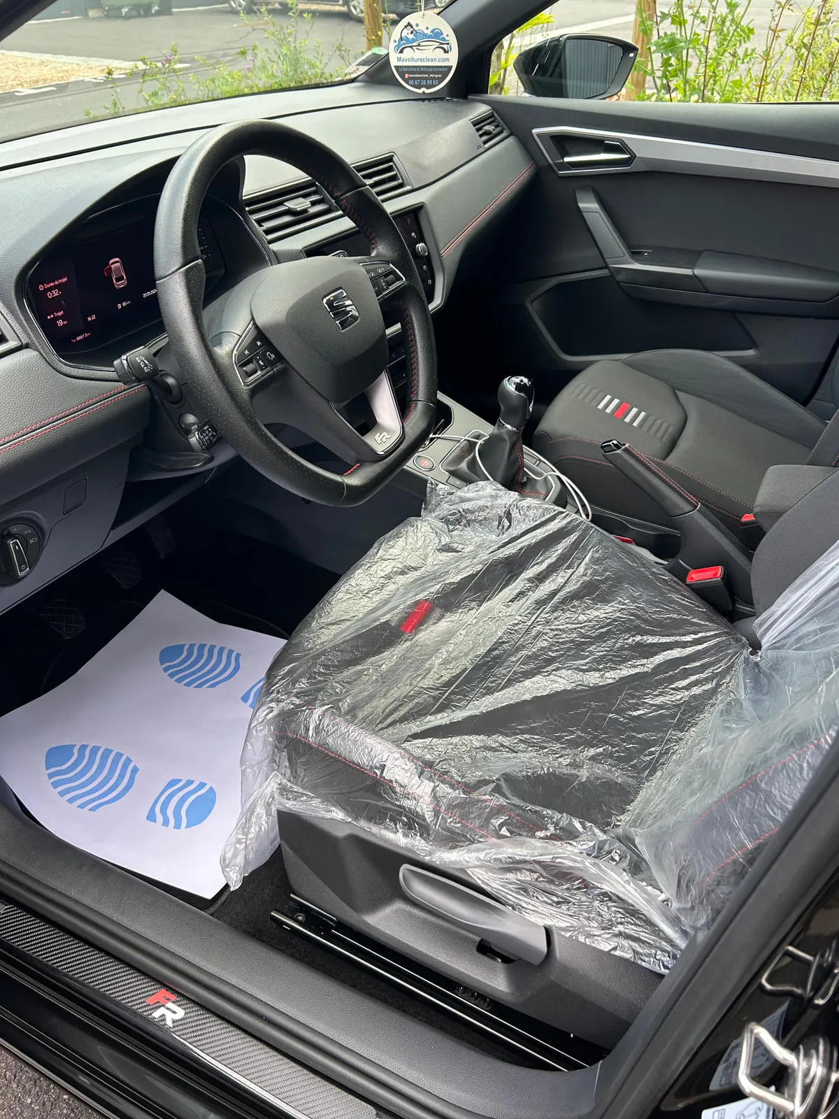 Poste de conduite d'une Seat vu par la portière ouverte : volant au logo Seat et à l'insigne FR, siège avant sous film plastique, feuille de protection à motif bleu posée sur le plancher, seuil de porte à motif carbone portant FR