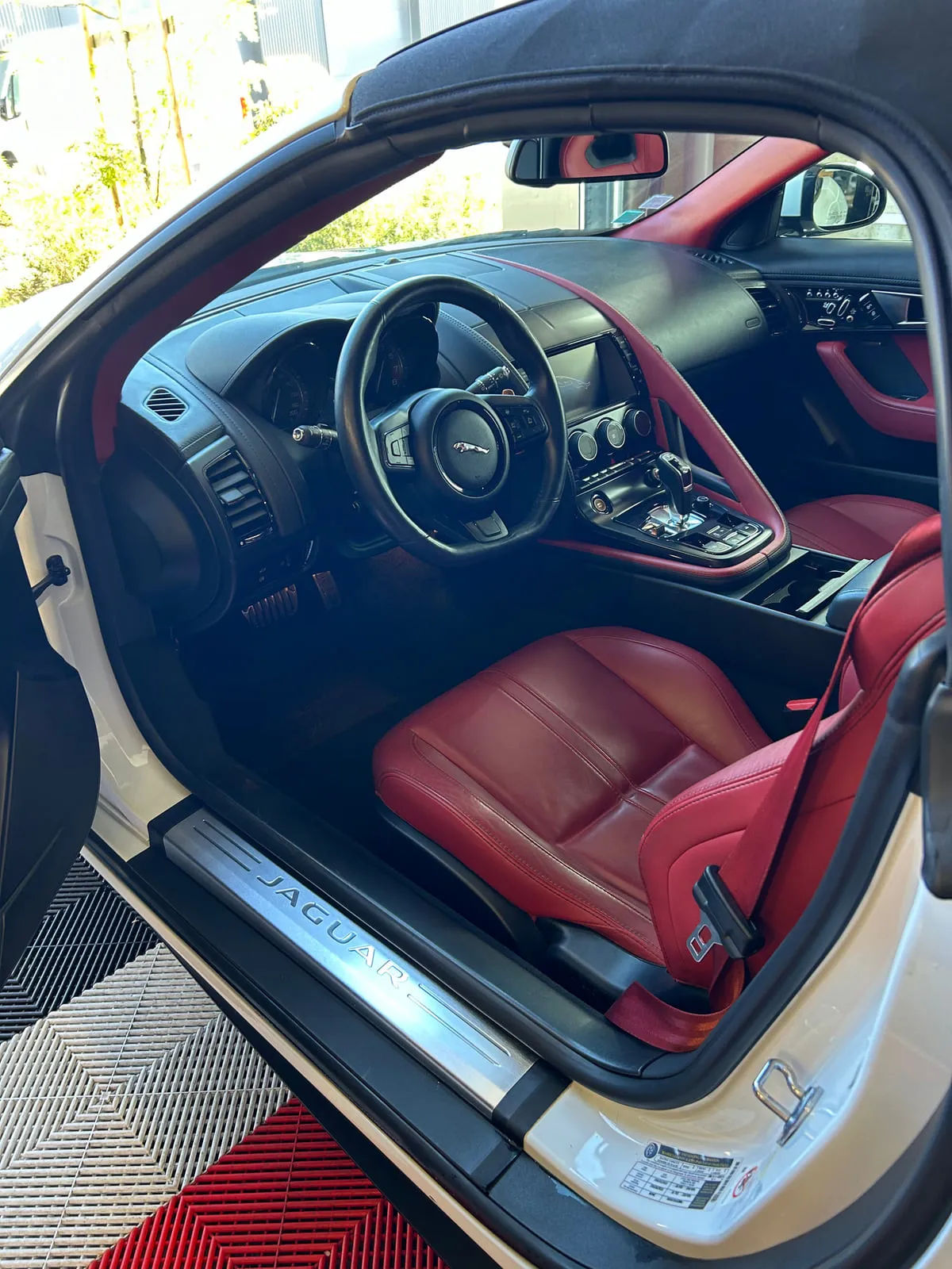 Siège avant en cuir rouge à surpiqûres, avec sa ceinture rouge, vu par la portière ouverte d'une voiture blanche ; seuil de porte en métal brossé marqué Jaguar